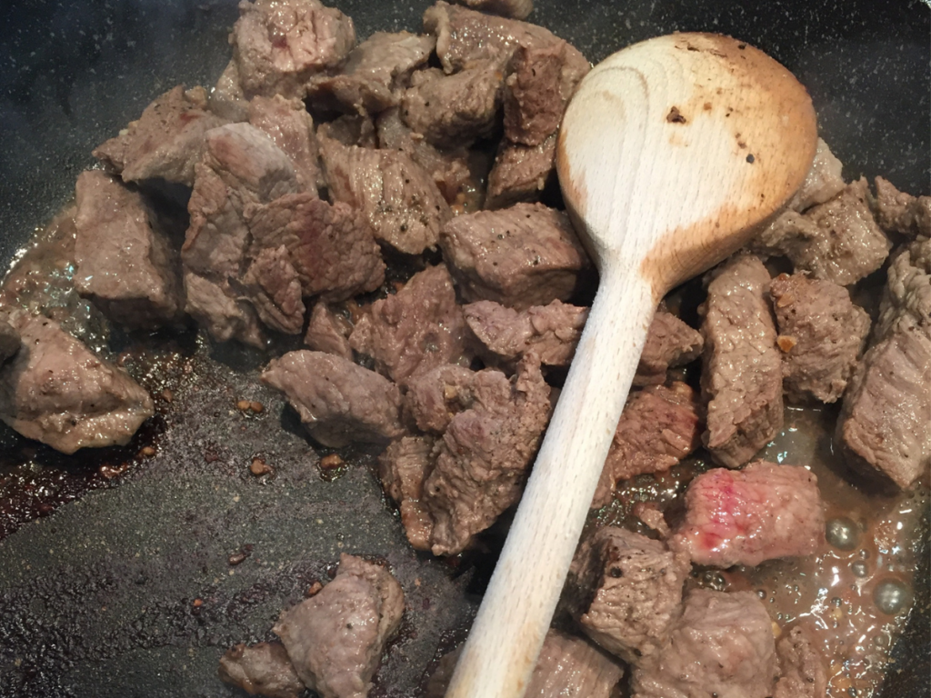 Cubes of beef are browning on a stovetop pan. The meat is cooking through though some pink flesh is still visible. It is being turned with a wooden spoon.