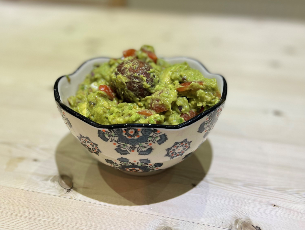 A bowl of fresh avocado guacamole containing pieces of tomato sits in a decorative bowl on a wooden table.