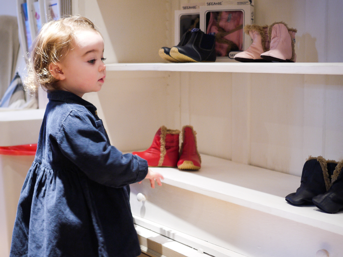 A small child of around 3 years old stands by a shoe display for little feet. She is in a large blue corduroy dress looking at the furry boots, by brand Skeanie.
