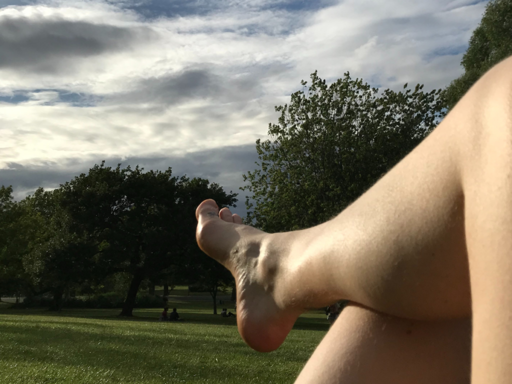 The leg and foot of a pale, white woman is reclined in a park. The sky is bright with dark clouds, and the trees are in full leaf. The grass is long and green. It is summer.