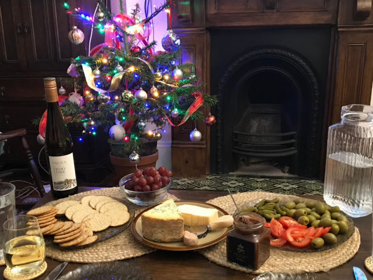 A coffee table is laid for a cheese course. On a plate, there are two large blocks of cheese. On another plate there are two types of biscuit for cheese. On a third plate are cornichons, olives and sliced tomatoes. A bottle of vinho verde is on the table. In the distance there is a small and brightly decorated Christmas tree,