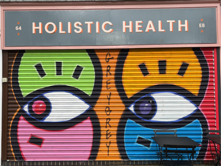 The shutter for the Holistic Health shop has been put down. It is brightly decorated by local artist Grey Grey, showing two large eyes looking to the side. The colours are green, pink, orange, yellow, blue and red.