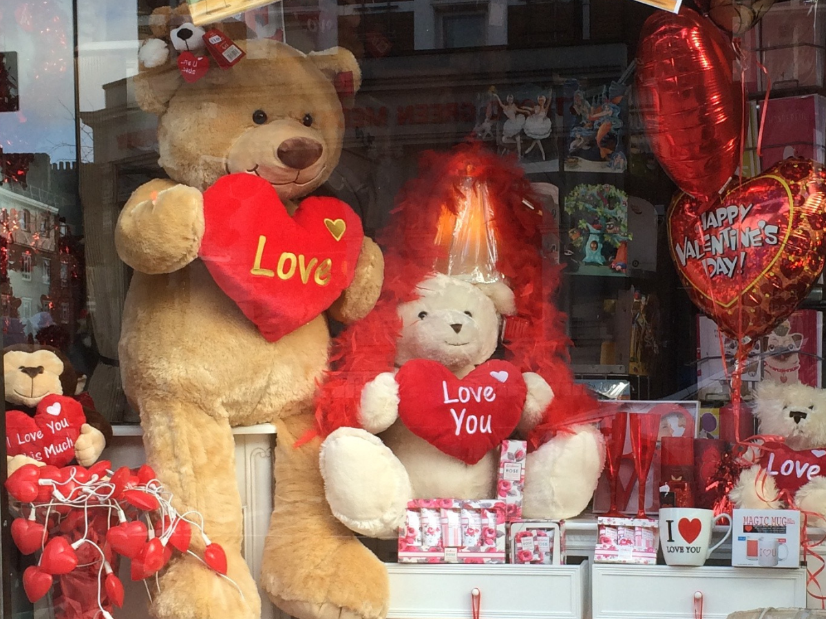 The window of a shop leading up to Valentines day. There are four teddy bears of varying sizes and species holding love hearts. There are balloons in the distance, mugs, and fairy lights, all covered in hearts.