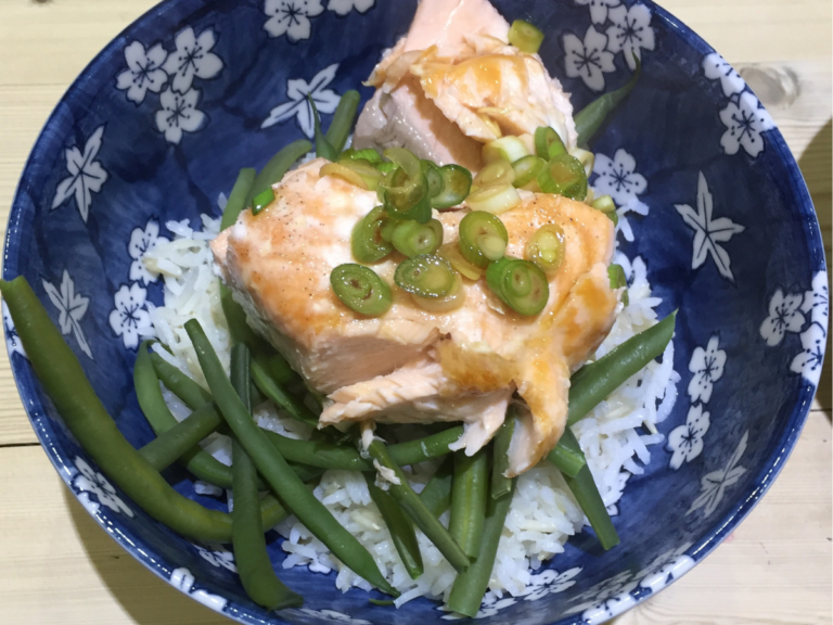 A navy blue bowl with white flowers on contains food. The food is a bed of white rice, green beans, and a fillet of cooked salmon topped with glistening spring onions.