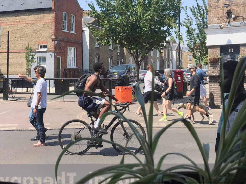 A view of Broadway Market taken from the clinic window. In the foreground a spider plant is visible. The people on the street are dressed for summer and the sun is shining. A cyclist and a greyhound are visible.