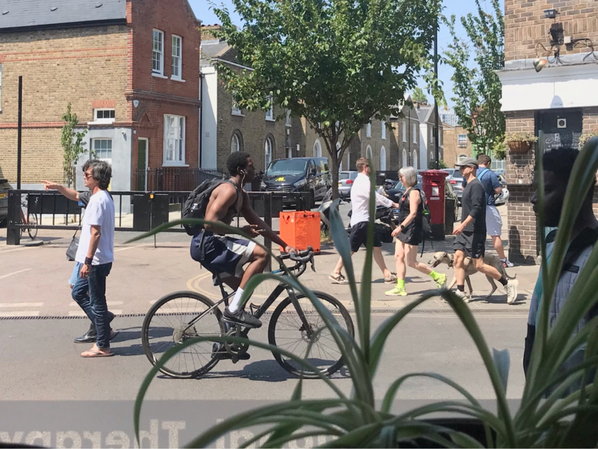 A view of Broadway Market taken from the clinic window. In the foreground a spider plant is visible. The people on the street are dressed for summer and the sun is shining. A cyclist and a greyhound are visible.