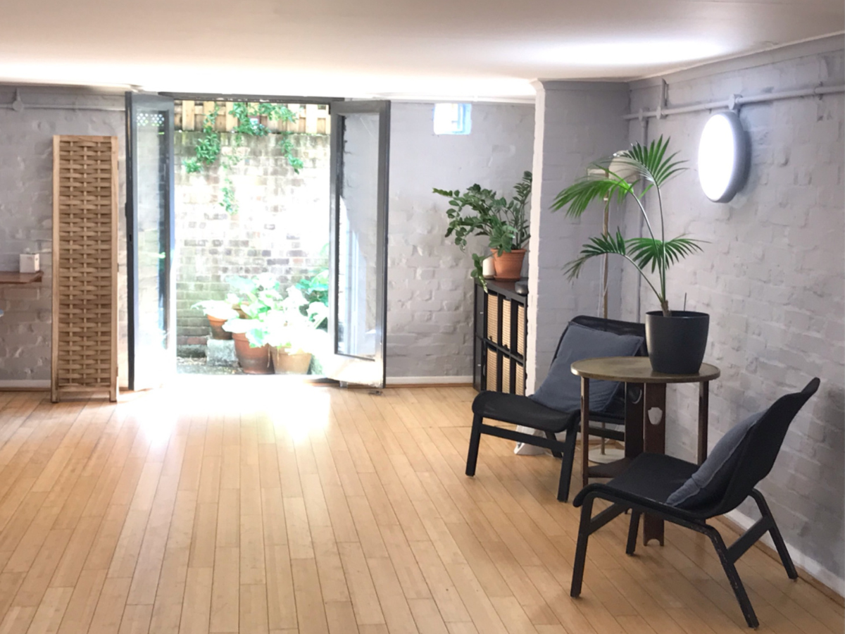 A landscape shot of the studio at Holistic Health. It is a large naturally lit room and the doors are open to the courtyard behind. To the right are two relaxed chairs and a golden vintage table. There are indoor plants on the surfaces.