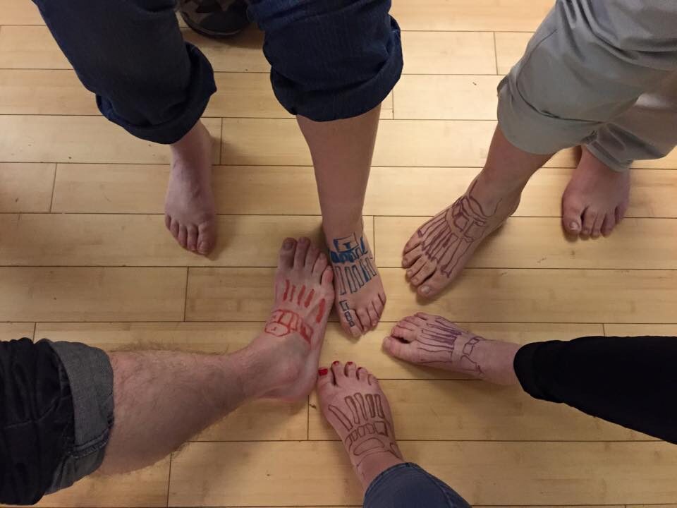 Five people have put their bare feet into the centre below the camera. They have drawn the metatarsal and tarsal bones of the foot, one person has drawn the phalanges of the big toe. They are likely completing an educational activity about feet.