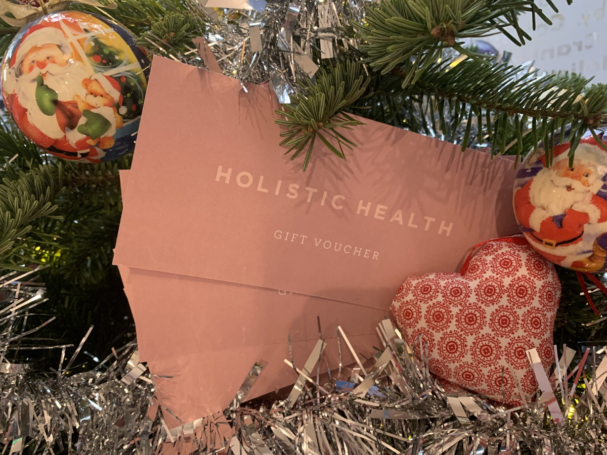 A close up photograph of two Holistic Health Gift Vouchers nestled amongst the branches of a tree. There are 2 Father Christmas baubles and a red and white fabric heart. Silver tinsel adorns the tree