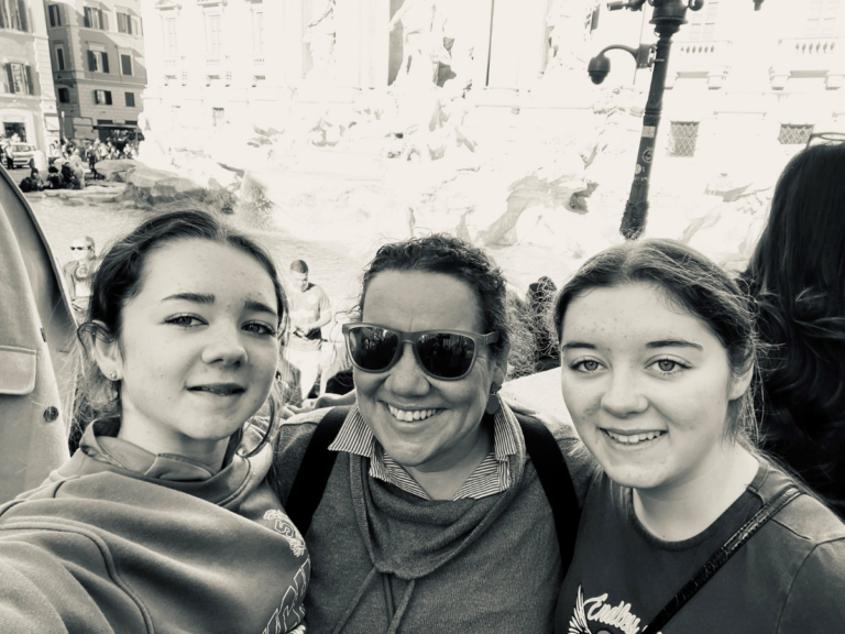 A black and white photo of Tracy with her daughters by the Treviso fountain in Rome. All 3 women are smiling. The sun is shining