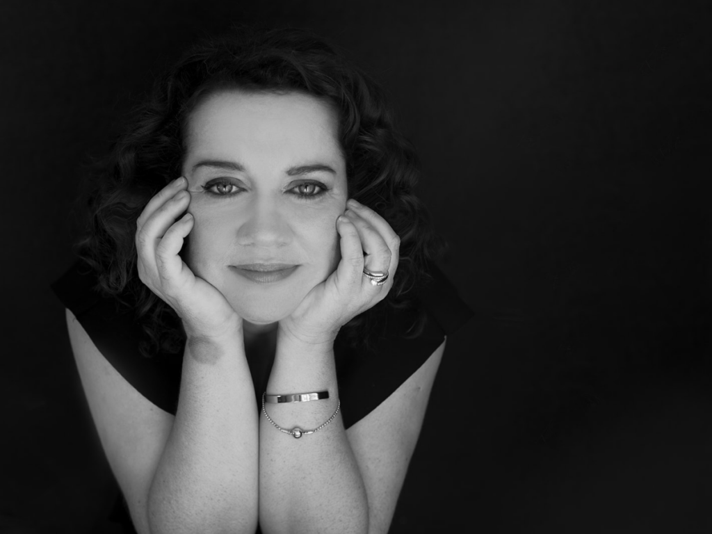 A black and white landscape portrait of Tracy Byrne, a smiling white woman. She wears a blacktop and silver jewellery. Her hair is dark and curly and she looks welcoming. She has a birthmark on her wrist.