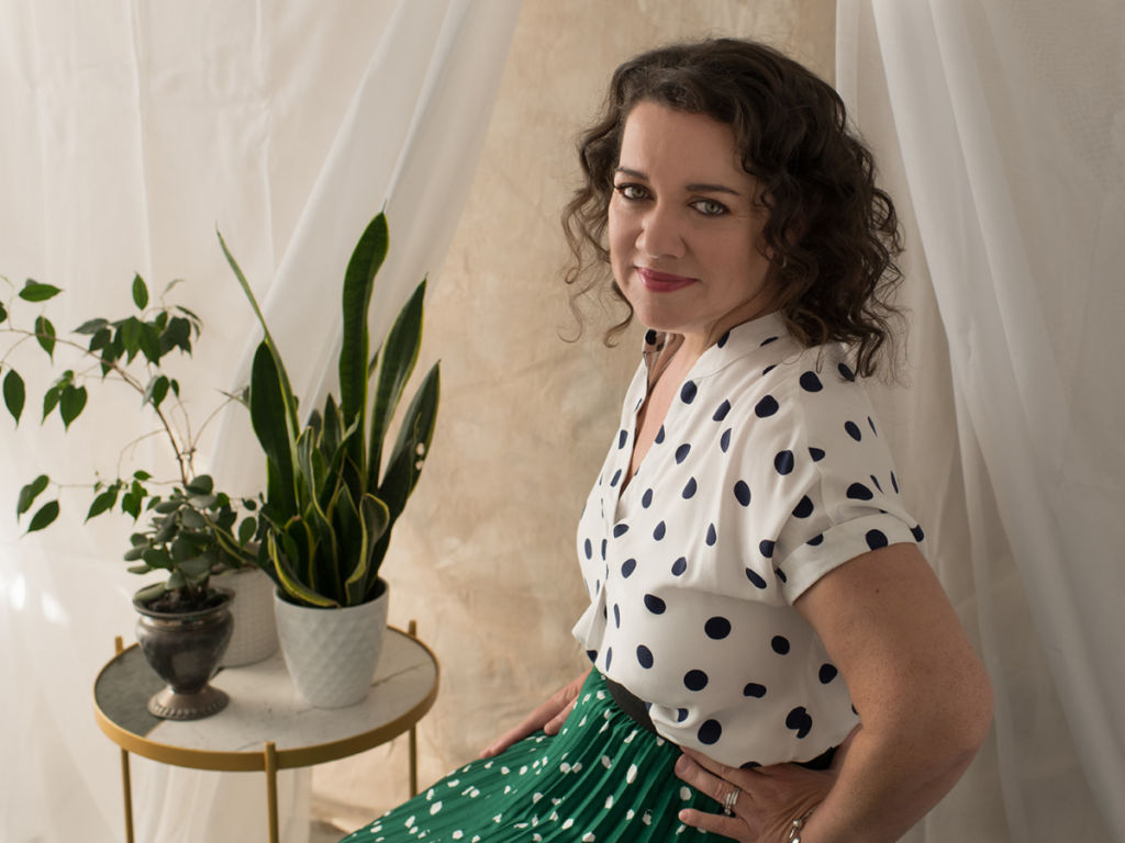 A landscape portrait of Tracy Byrne, a smiling white woman. She wears a black on white polka dot shirt. and a white on green polka dot skirt. Her hair is dark and curly and she looks welcoming. Plants are visible in the backgroun upon a circular table.