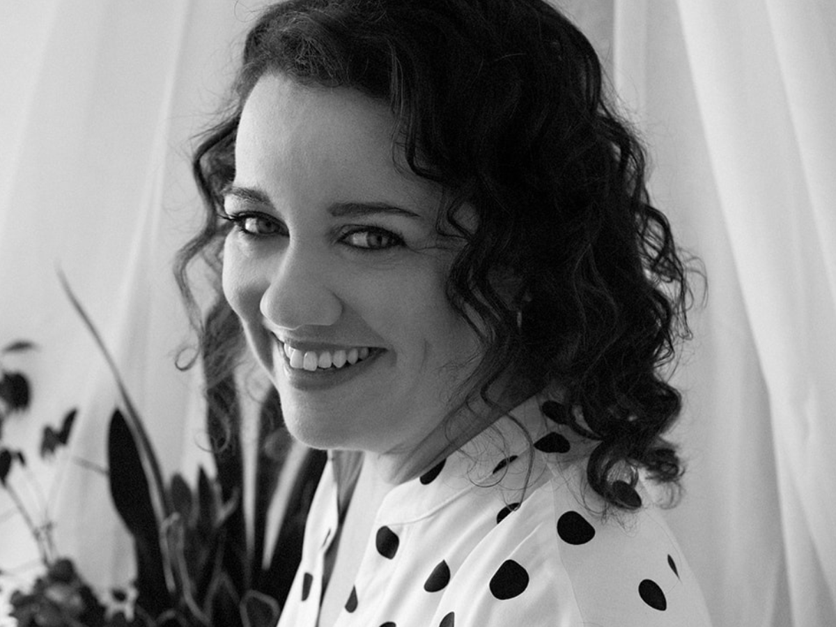 A black and white landscape portrait of Tracy Byrne, a smiling white woman. She wears a black on white polka dot shirt. Her hair is dark and curly and she looks welcoming. Plants are visible in the background.